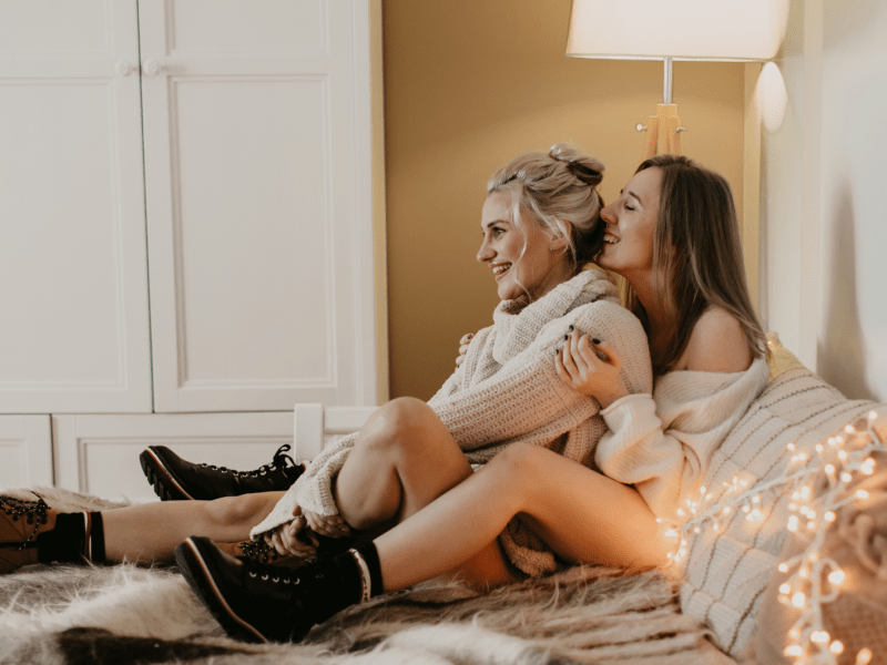 Zwei junge Frauen in Strickpullovern sitzen lachend auf einem Fellteppich, umgeben von Lichterketten, in einem gemütlich beleuchteten Zimmer.