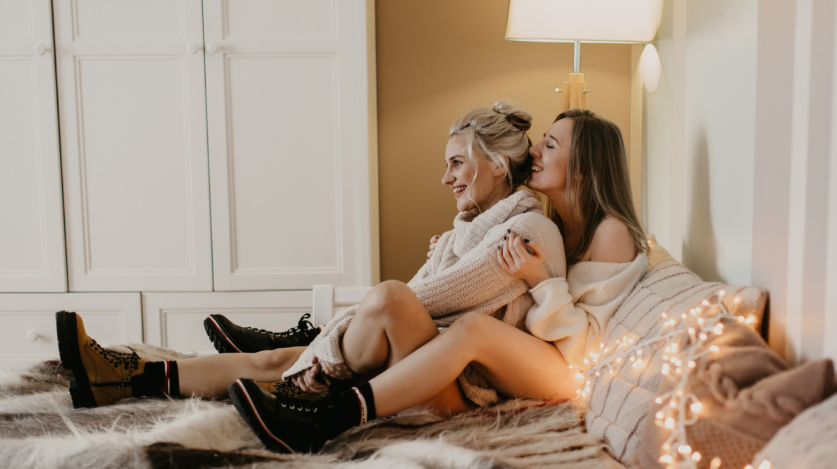 Zwei junge Frauen in Strickpullovern sitzen lachend auf einem Fellteppich, umgeben von Lichterketten, in einem gemütlich beleuchteten Zimmer.