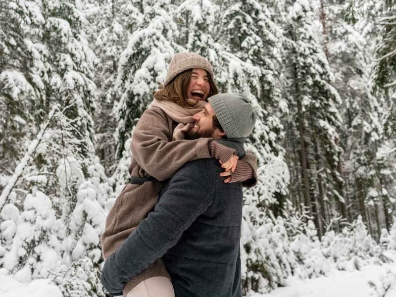 Mann hebt fröhliche Frau im braunen Mantel hoch, beide lachen herzlich in einem verschneiten Nadelwald.