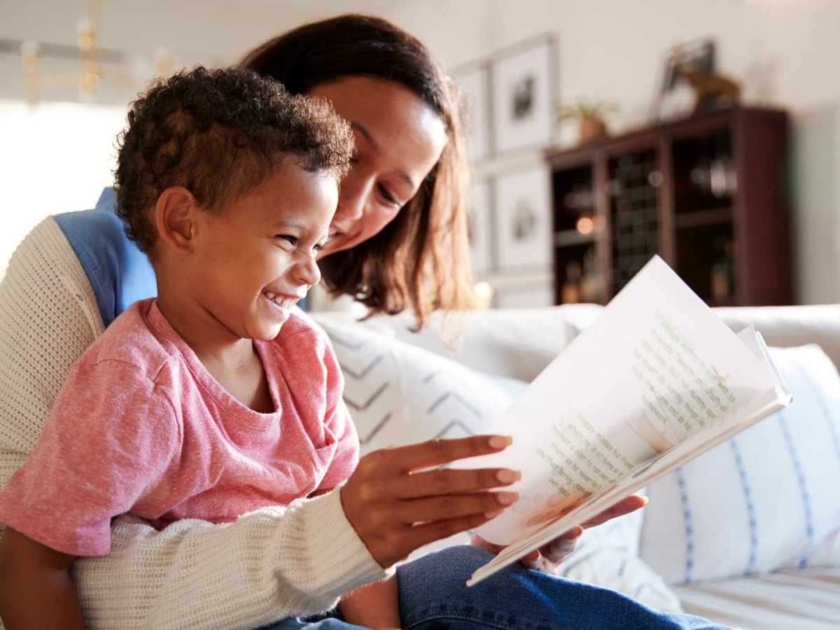 Mutter und Sohn, ca. 4 Jahre alt, sitzen auf dem Sofa und lesen gemeinsam ein Buch. Das Kind lacht.