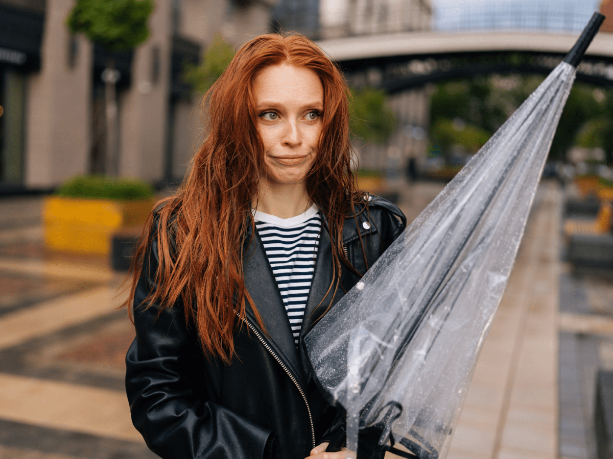 Frau mit nassen und durchwuschelten Haaren, die einen Regenschirm in der Hand h&auml;lt und schmollend zur Seite schaut.