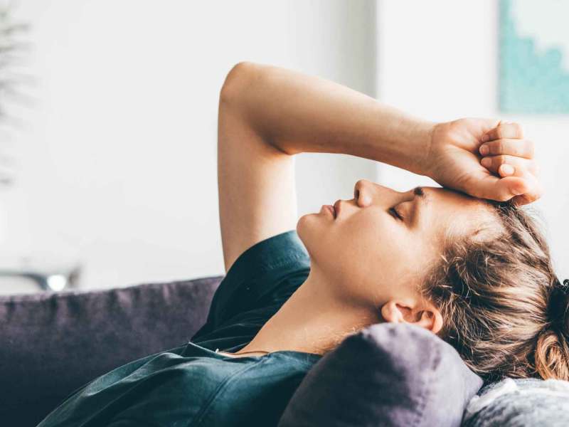 Eine junge Frau mit dunklem Dutt lehnt erschöpft auf einem Sofa, ihre Augen geschlossen und die Hand an die Stirn gelegt.