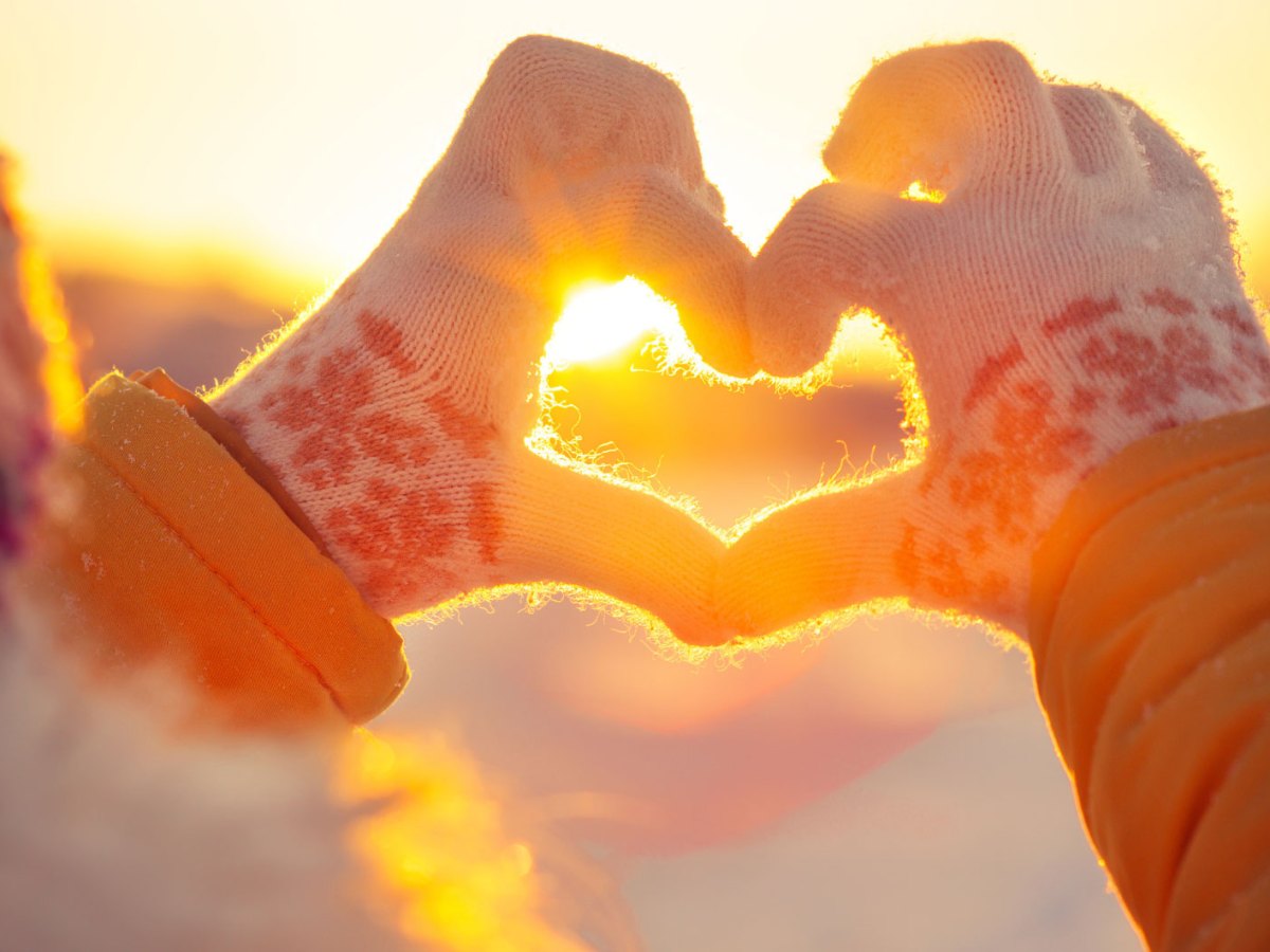 Person in Winterbekleidung formt ein Herz mit ihren H&auml;nden in winterlicher Landschaft bei Sonnenuntergang