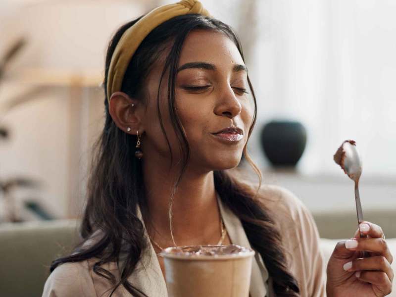 Junge Frau mit dunklen, welligen Haaren und gelbem Haarband genießt mit geschlossenen Augen Schokoladendessert auf einem Löffel.