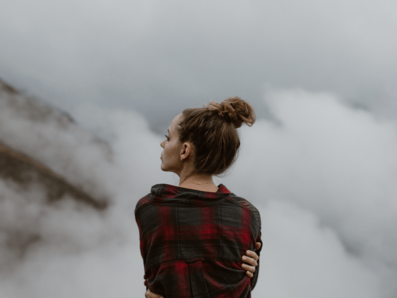 Frau mit Dutt auf einem Berg im Nebel, die die Arme um sich schlingt und nachdenklich zur Seite blickt.