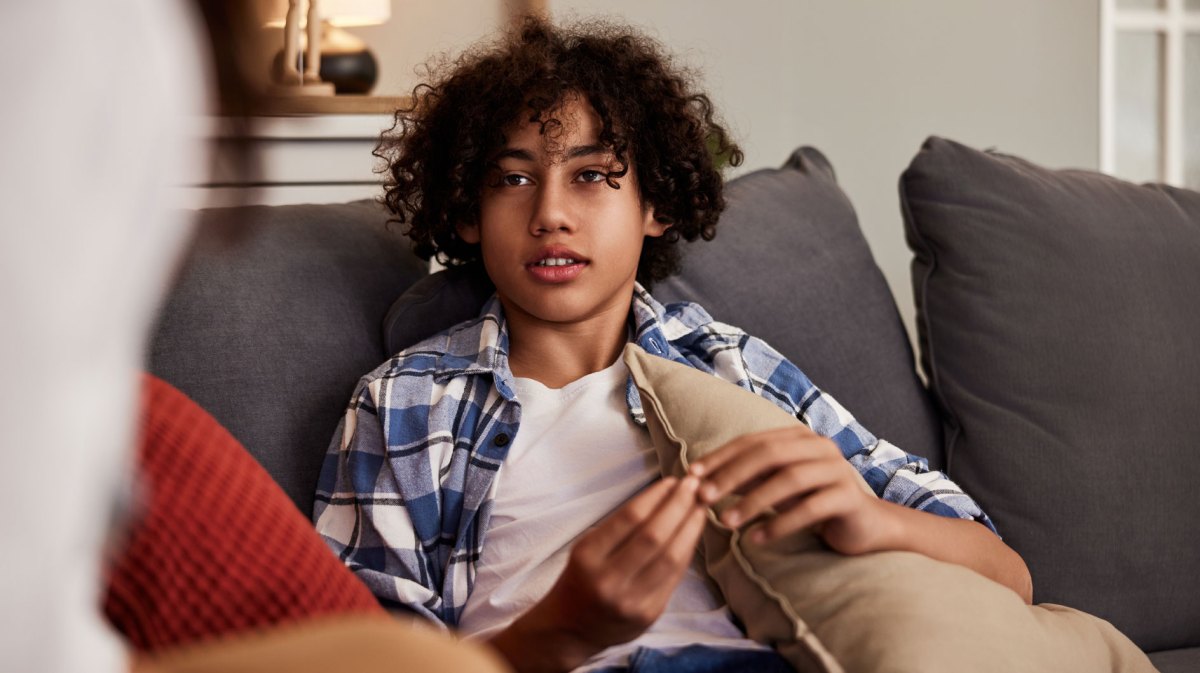Porträt eines Teenagers, der auf einer Couch sitzt, ein Kissen festhält und ernst mit jemandem spricht.