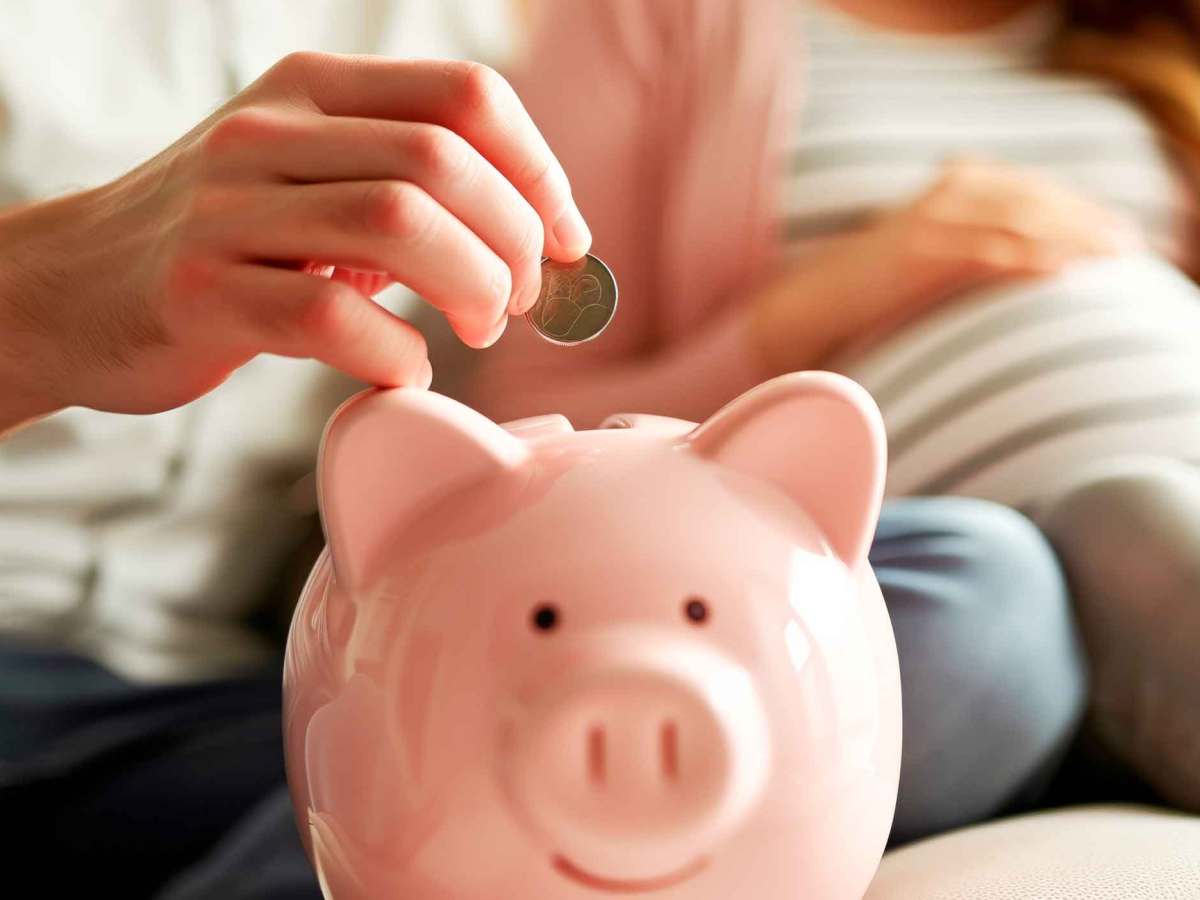 Mann wirft eine M&uuml;nze in ein rosa Sparschwein. Im Hintergrund sitzt eine schwangere Frau.