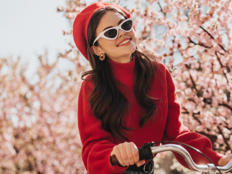 Frau mit rotem Pulli und Barett fährt Fahrrad, im Hintergrund sind Kirschblüten zu sehen.