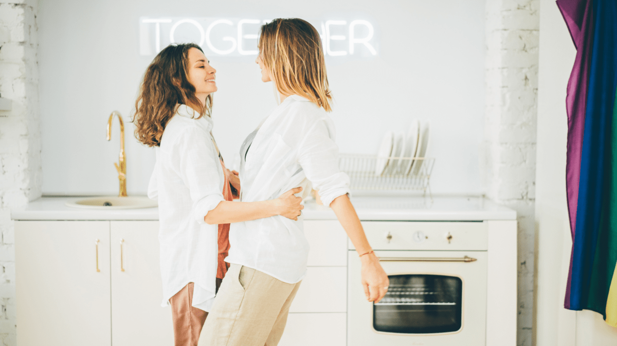 Paar hat Spaß in Küche mit Regenbogenflagge. Frauen tanzen und drehen, genießen die Zweisamkeit auf Hintergrund Neonschild mit Wort Together. Konzept LGBT