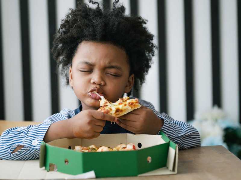 Glücklihces Kind beißt genussvoll und mit geschlossenen Augen in ein Stück Pizza. Vor ihm auf dem Tisch steht der Pizzakarton.