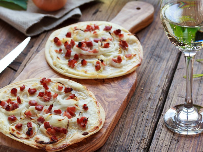 Zwei kleine runde Flammkuchen auf Holzbrett auf Tisch.