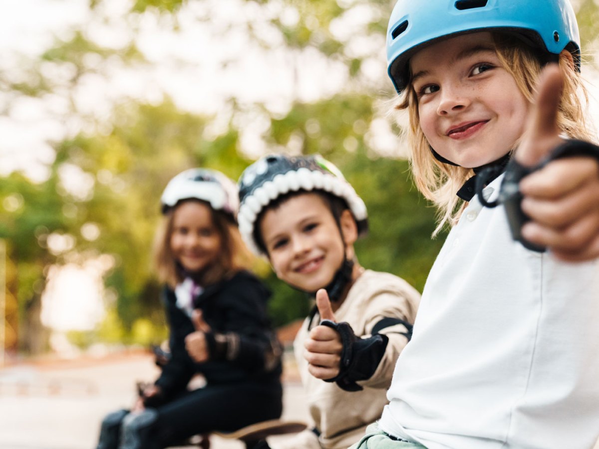 Drei Kinder sitzen in voller Sicherheitsmontur auf dem Skaterplatz und strecken den Daumen hoch Richtung Kamera.