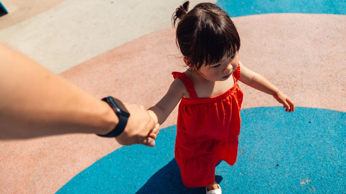 Mädchen in rotem Kleid an Hand von Erwachsenem als Symbolbild für Helikopter-Eltern