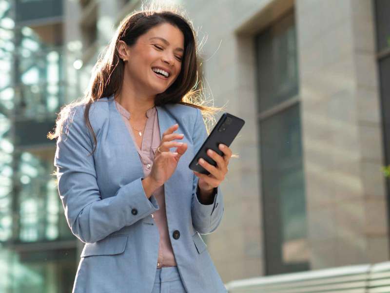 Junge Frau mit schulterlangem braunem Haar und hellblauem Anzug lächelt und tippt auf ihr Smartphone in sonniger Büroumgebung.
