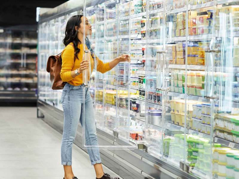 Eine junge Frau mit langen schwarzen Haaren und gelbem Pullover greift in einem Supermarkt nach einem Produkt aus einer Kühltheke.