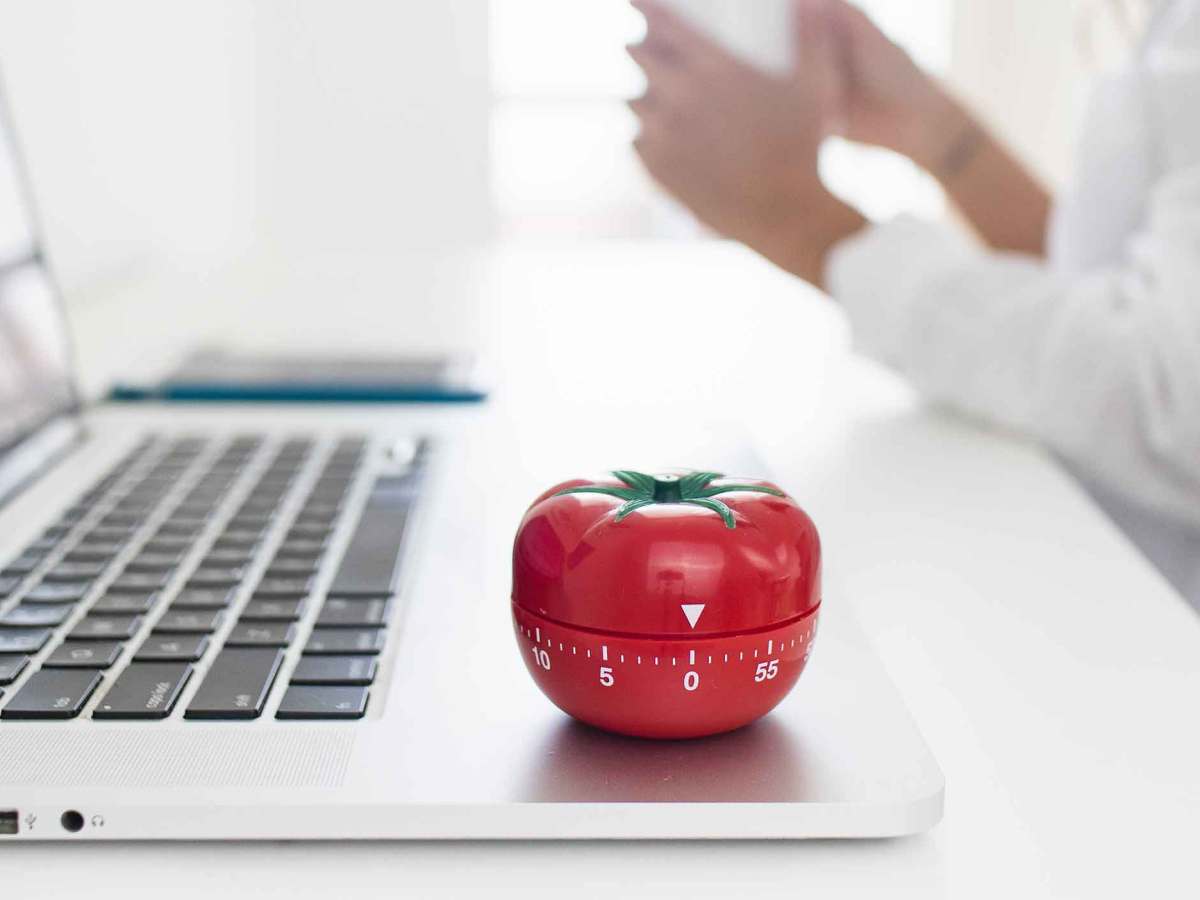 Eine K&uuml;chenuhr in Form einer Tomate steht auf einem Notebook. Im Hintergrund sitzt eine Frau mit einem Kaffee.