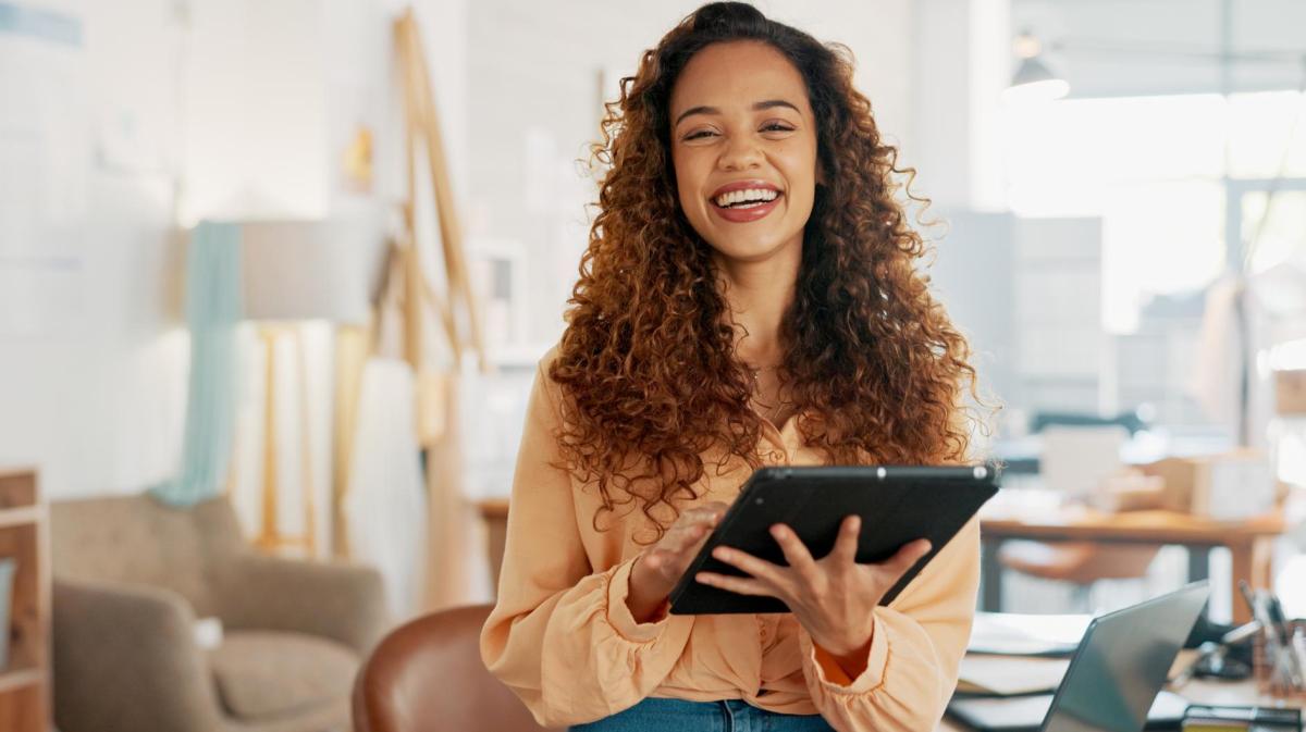 Frau mit Tablet steht im Büro und lacht.