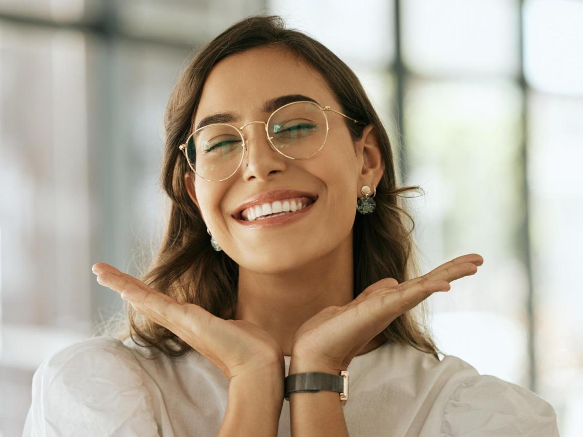 Junge Frau mit Brille umrahmt ihr Gesicht mit ihren H&auml;nden und grinst.