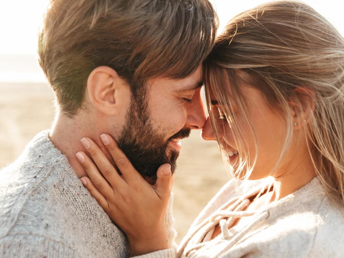 Ein verliebtes junges Paar in hellen Strickpullovern steht eng umschlungen am Strand, Stirn an Stirn, und lächelt sich an.