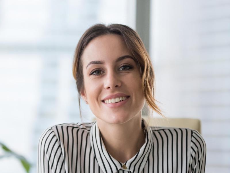 Junge Frau mit zusammengesteckten Haaren und einem gestreiften Hemd lächelt am Schreibtisch.