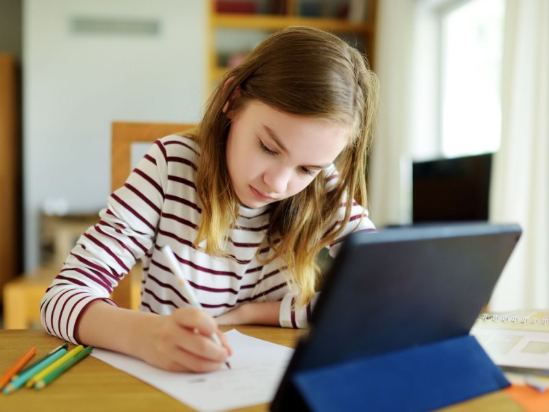 Mädchen, ca. 9 Jahre alt, sitzt mit einem Tablet, Papier und Stiften am Tisch und erledigt Aufgaben.