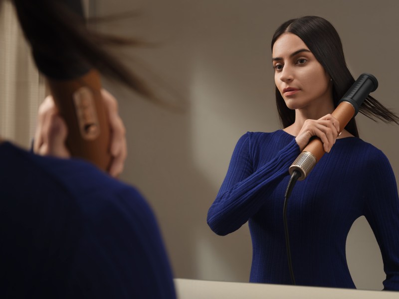 Brünette Frau im blauen Pullover glättet ihr Haar mit einem Haarstylinggerät vor einem Spiegel, ihr Blick ist fokussiert.