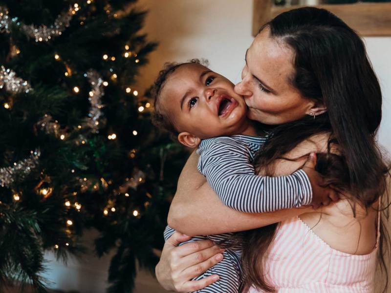 Mutter gibt ihrem weinenden Baby einen Kuss auf die Wange, w&auml;hrend beide vor dem Weihnachtsbaum sitzen.