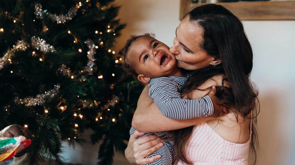 Mutter gibt ihrem weinenden Baby einen Kuss auf die Wange, während beide vor dem Weihnachtsbaum sitzen.