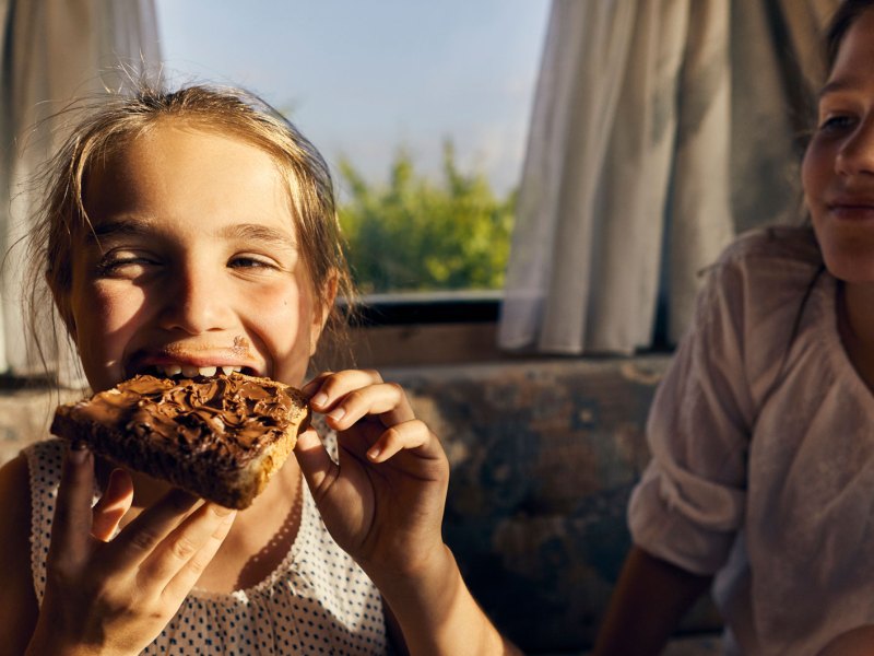 Zwei Schwestern, eine isst ein Nutellabrot, die andere schaut neidisch zu.