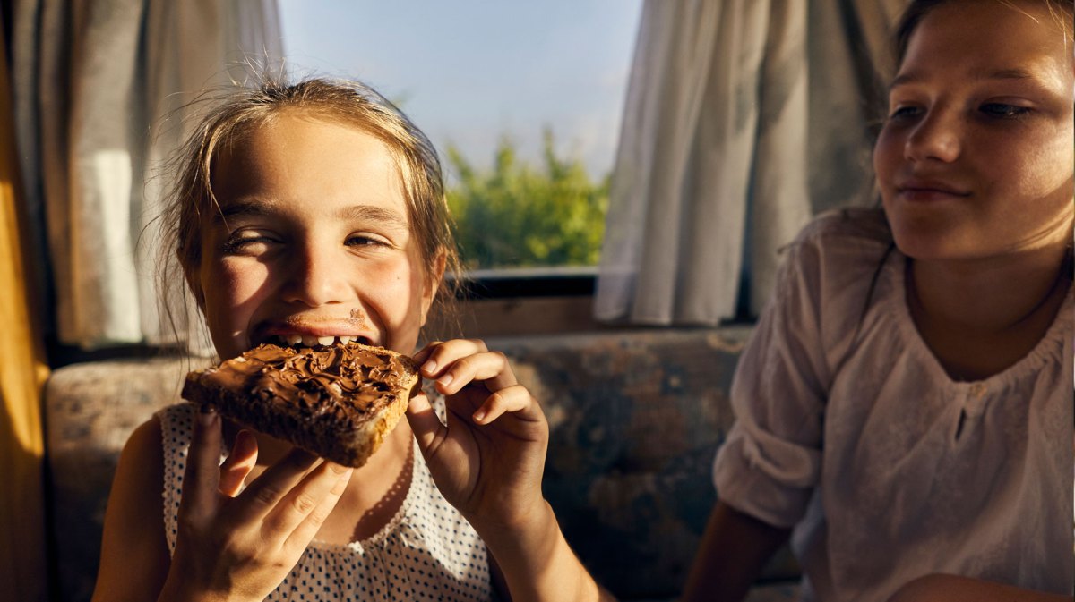 Zwei Schwestern, eine isst ein Nutellabrot, die andere schaut neidisch zu.