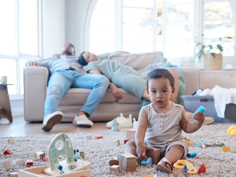 Schlafende Eltern liegen auf dem Sofa. Davor sind viele Spielsachen und ein Kleinkind