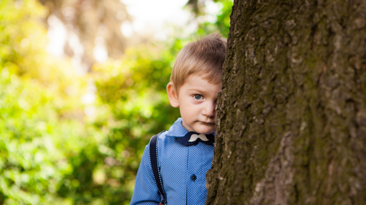 Junge, etwa 7 Jahre alt, steht schüchtern und halb versteckt hinter einem Baum