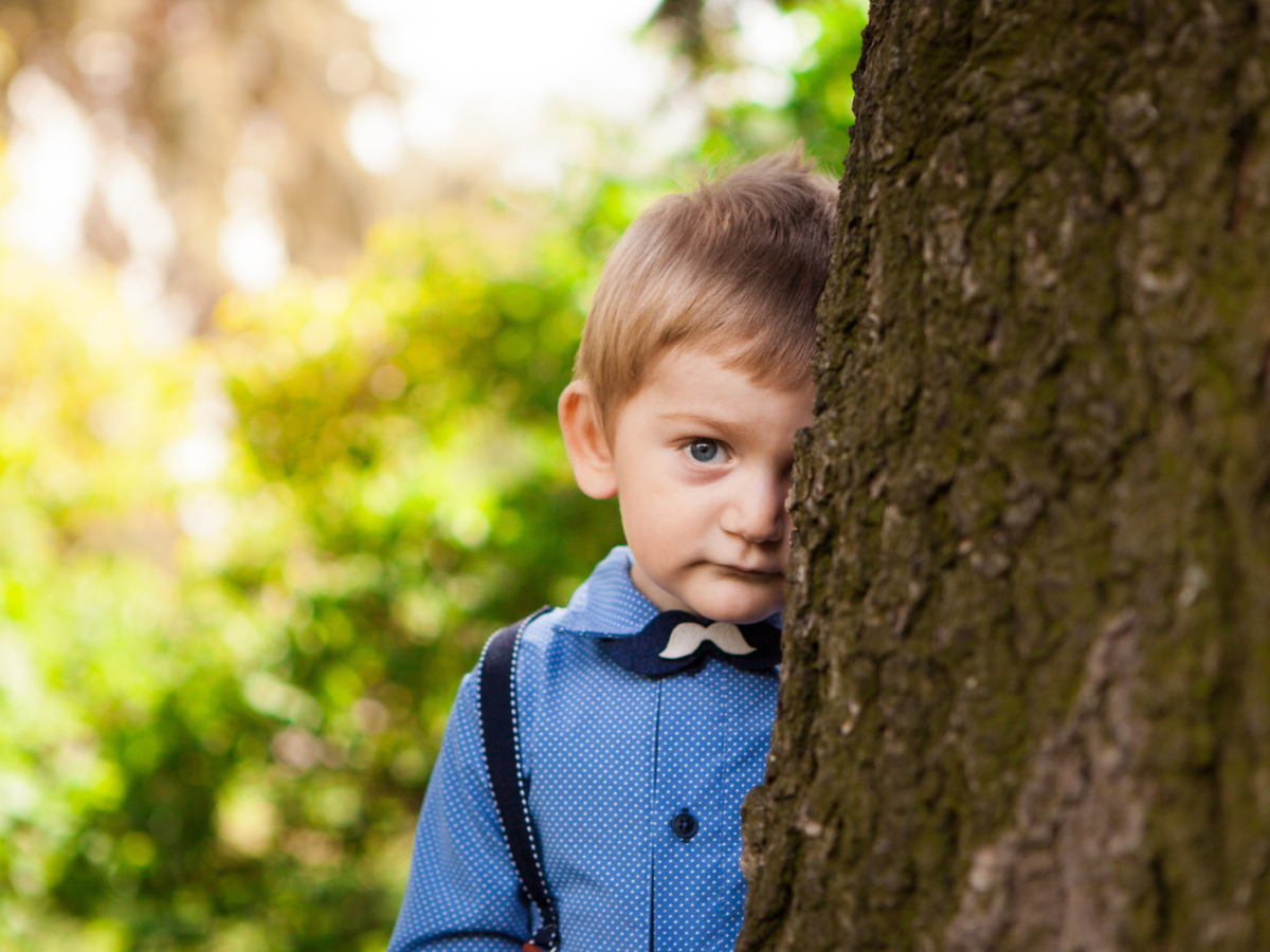 Junge, etwa 7 Jahre alt, steht schüchtern und halb versteckt hinter einem Baum