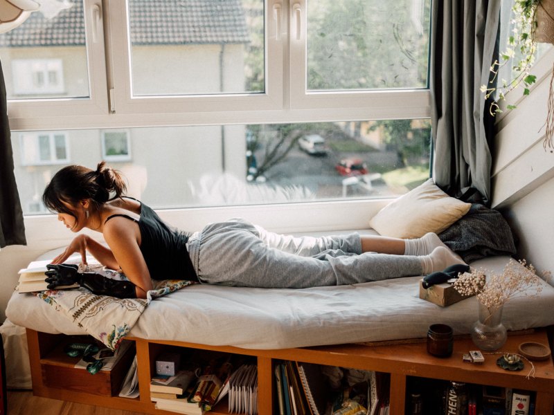 Junge Frau liegt auf einer Matratze am Fenster und liest etwas.