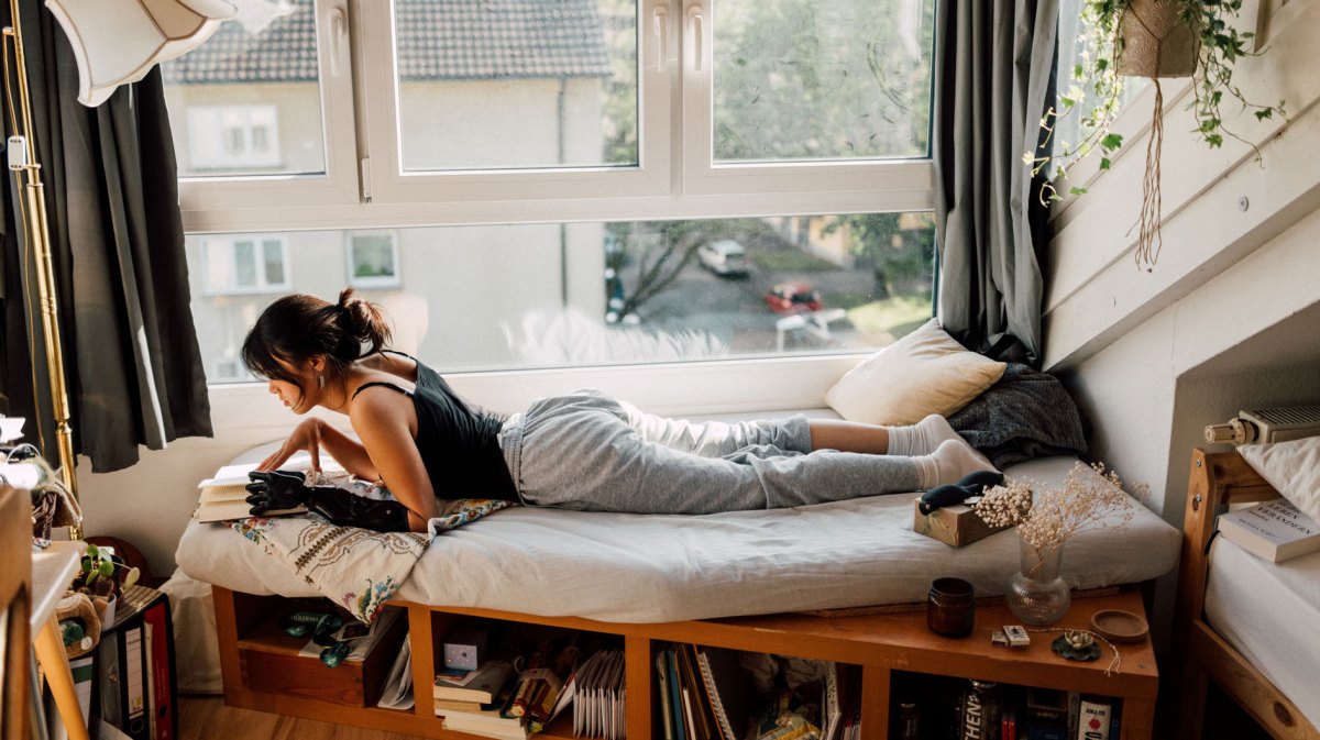 Junge Frau liegt auf einer Matratze am Fenster und liest etwas.