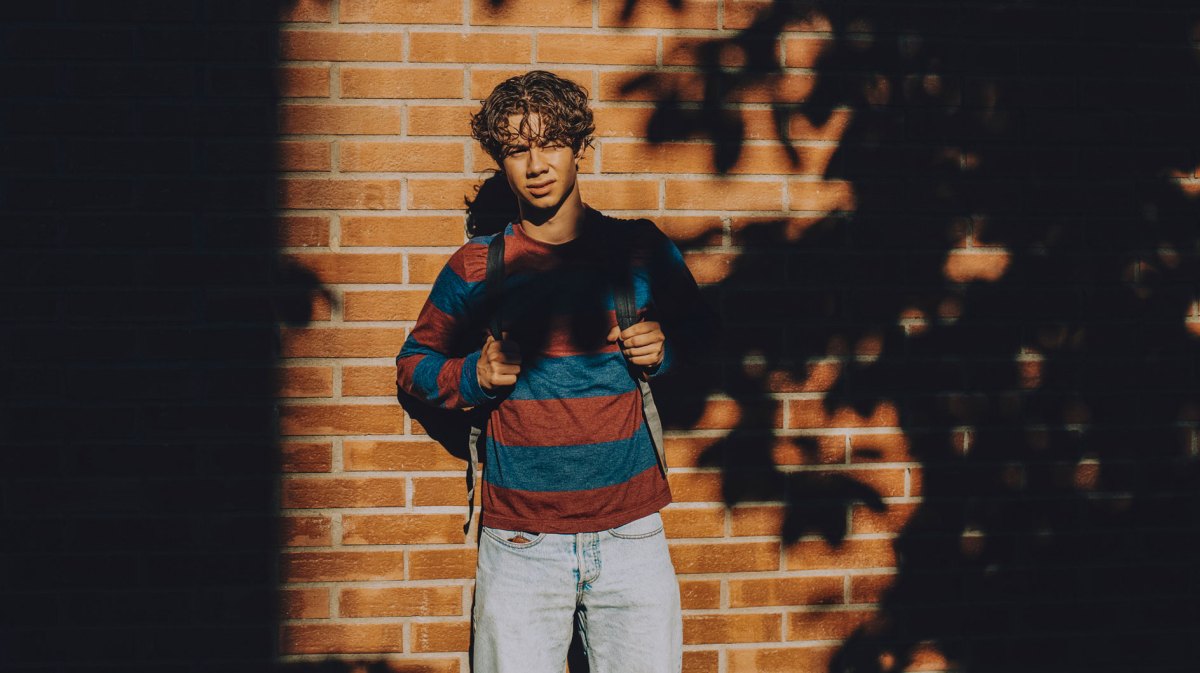 Junge im Teenageralter steht vor einer gemauerten Wand im Halbschatten und schaut ernst in Richtung Kamera. Er trägt Jeans, Sweatshirt und Rucksack.