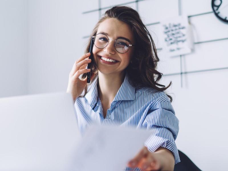 Junge Frau sitzt am Schreibtisch mit Brille und telefoniert.