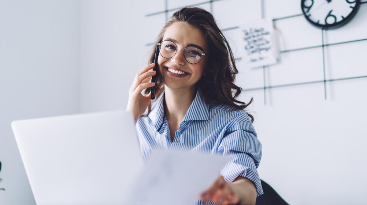 Junge Frau sitzt am Schreibtisch mit Brille und telefoniert.
