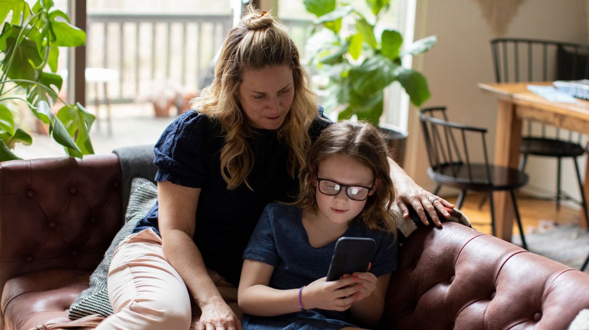 Mutter sitzt mit ihrer Tochter, ca. 8 Jahre alt, auf der Couch und schaut gemeinsam mit ihr aufs Smartphone.
