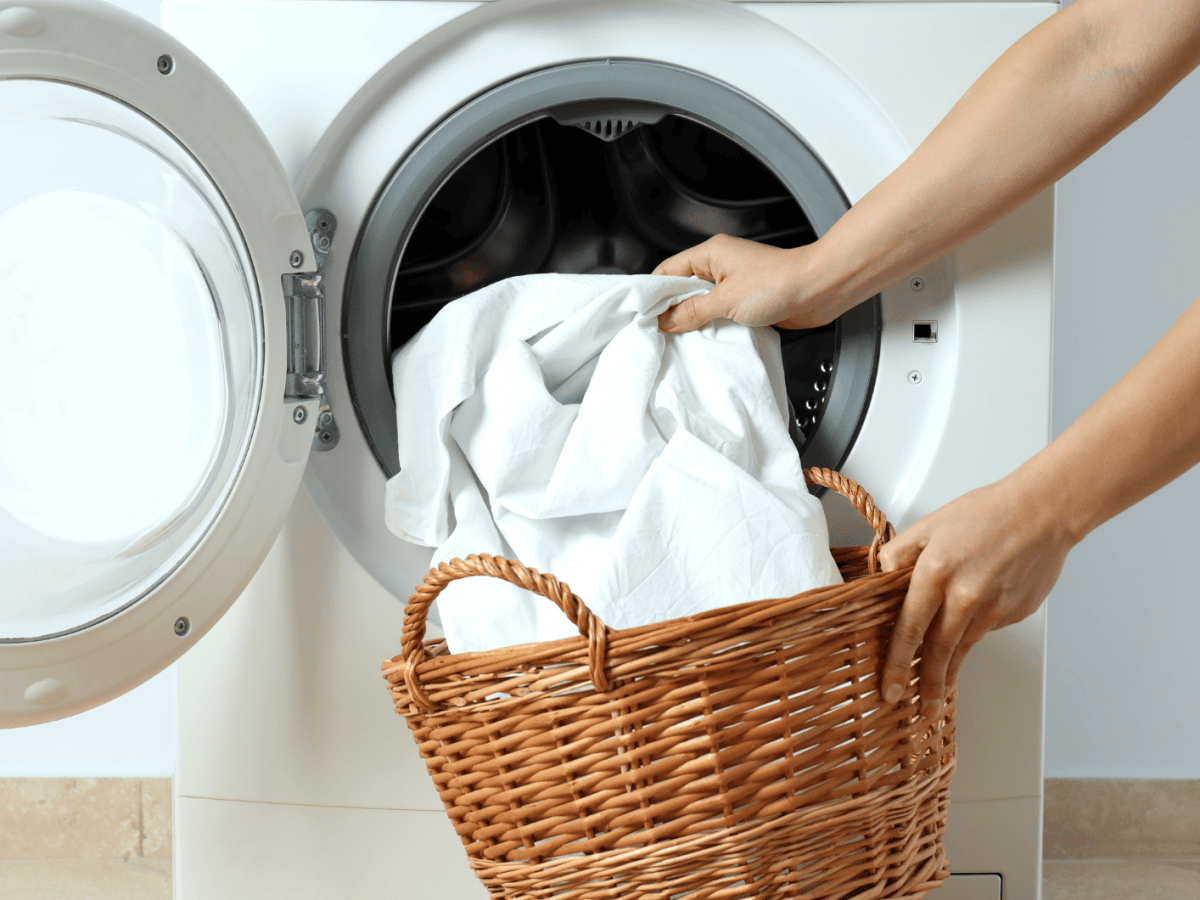 Person gibt weiße Wäsche aus einem Strohkorb in die Waschmaschine