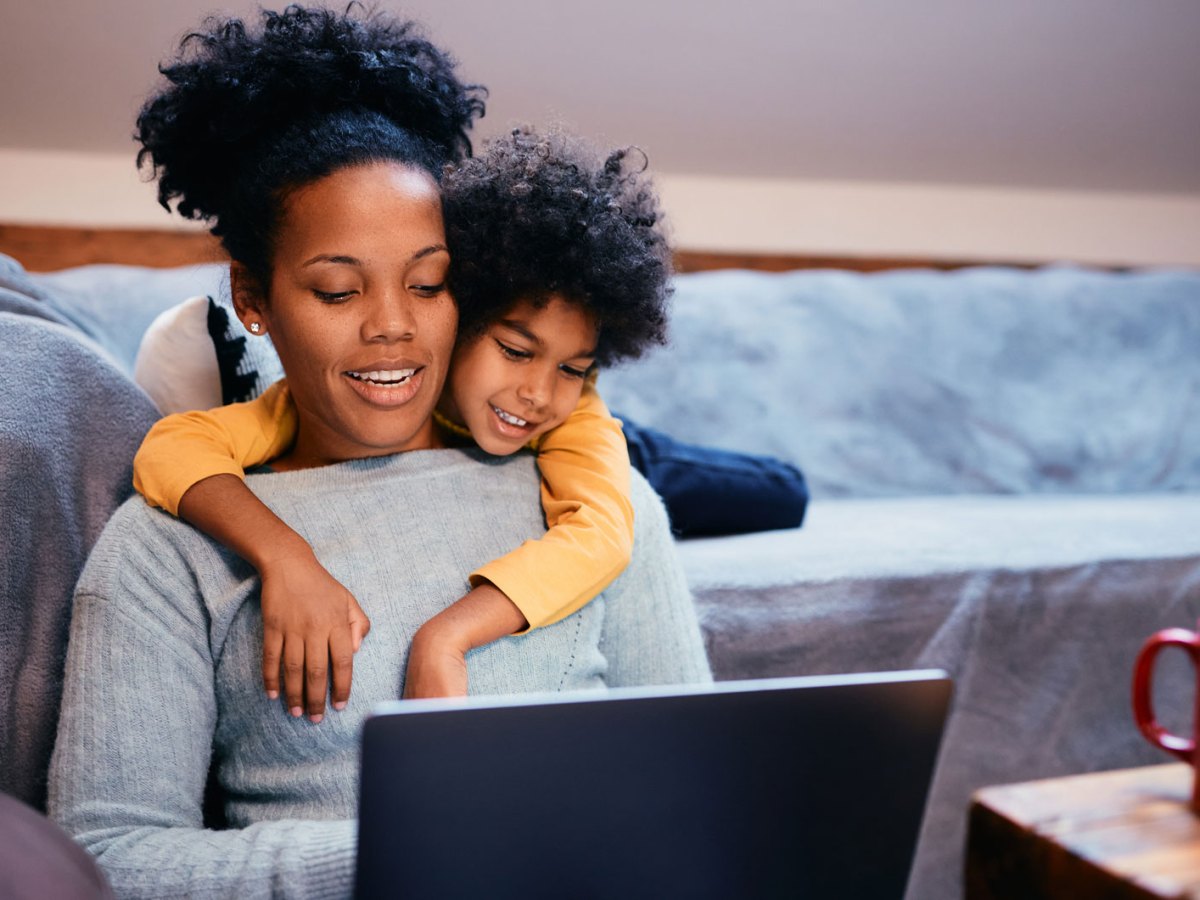 Mutter und Tochter sitzen im Wohnzimmer und schauen etwas am Laptop. Die Mutter sitzt auf dem Fußboden, an die Couch gelehnt, die Tochter schlägt von hinten ihre Arme um die Mutter und schaut ihr über die Schulter.
