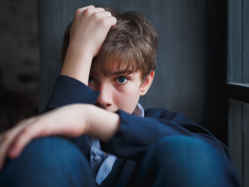 Junge, ca. 13 Jahre, sitzt zusammengekauert am Fenster und versteckt sein Gesicht zur Hälfte hinter seien Armen. Das Bild ist insgesamt in dunklen Farben gehalten.