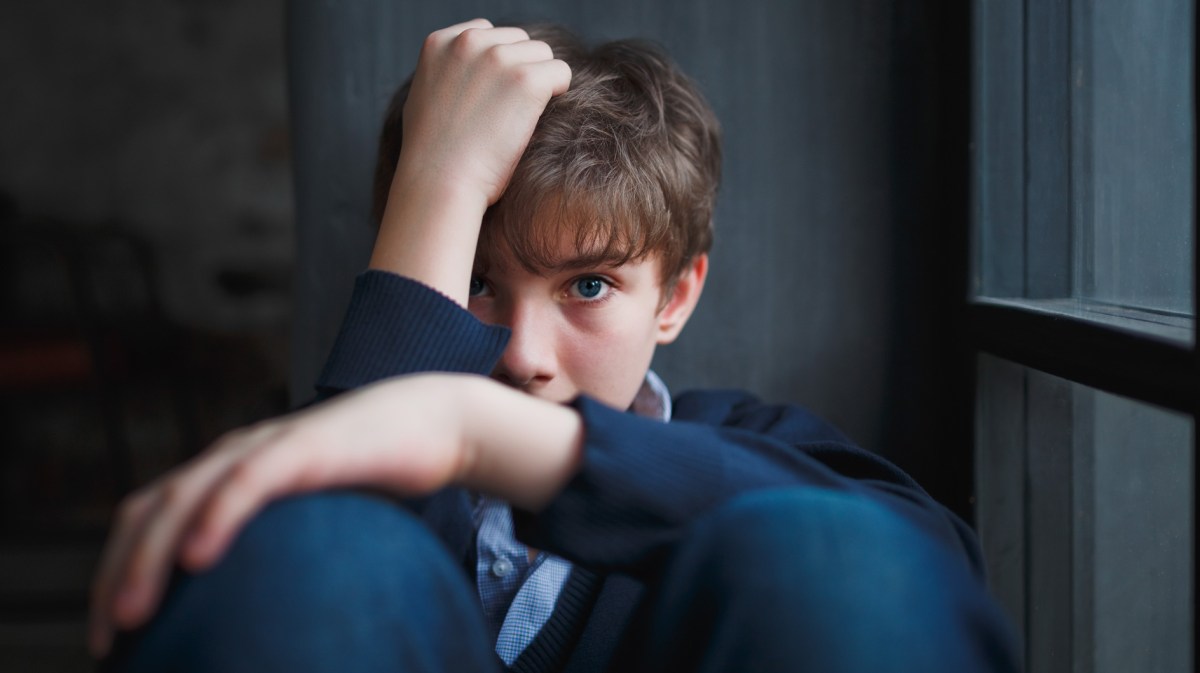 Junge, ca. 13 Jahre, sitzt zusammengekauert am Fenster und versteckt sein Gesicht zur Hälfte hinter seien Armen. Das Bild ist insgesamt in dunklen Farben gehalten.