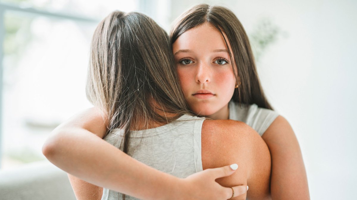 Besorgtes Teenager-Mädchen umarmt ihre Mutter. Das Mädchen schaut dabei direkt in die Kamera, die Mutter ist nur von hinten zu sehen.