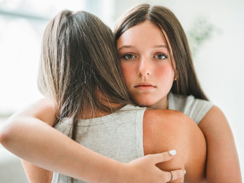 Besorgtes Teenager-Mädchen umarmt ihre Mutter. Das Mädchen schaut dabei direkt in die Kamera, die Mutter ist nur von hinten zu sehen.