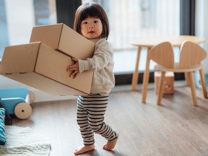 Kleinkind tr&auml;gt zwei gro&szlig;e Pappkartons durch ein Zimmer.