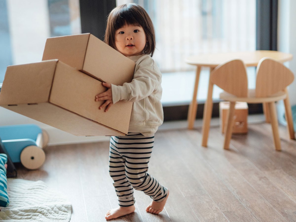 Kleinkind tr&auml;gt zwei gro&szlig;e Pappkartons durch ein Zimmer.