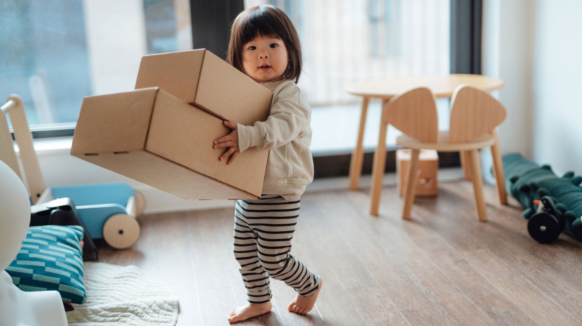Kleinkind trägt zwei große Pappkartons durch ein Zimmer.
