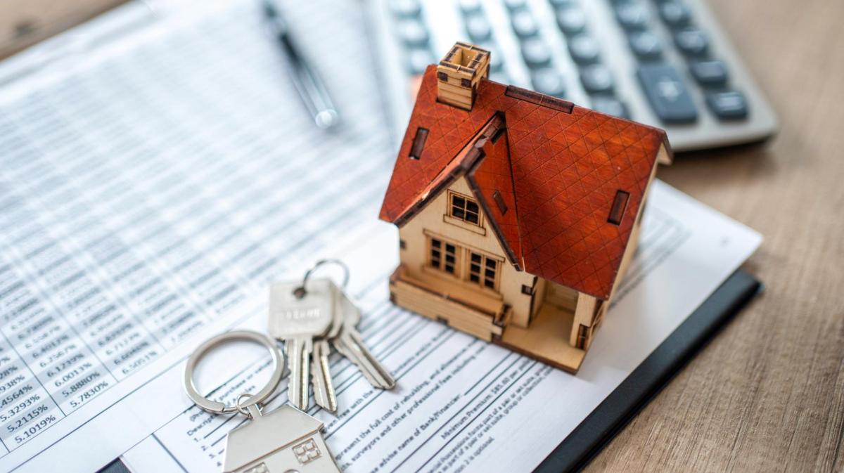 Miniaturhaus mit rotem Dach steht auf Vertragsdokumenten neben Hausschlüsseln, Kugelschreiber und Taschenrechner.