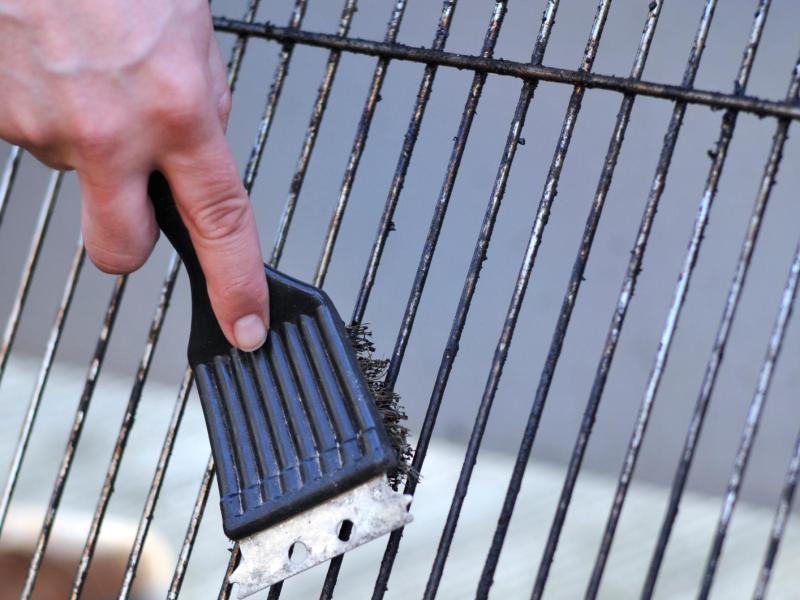 Eine Hand mit Grillbürste reinigt einen stark verschmutzten Grillrost über hellem Boden im Freien bei Tageslicht.